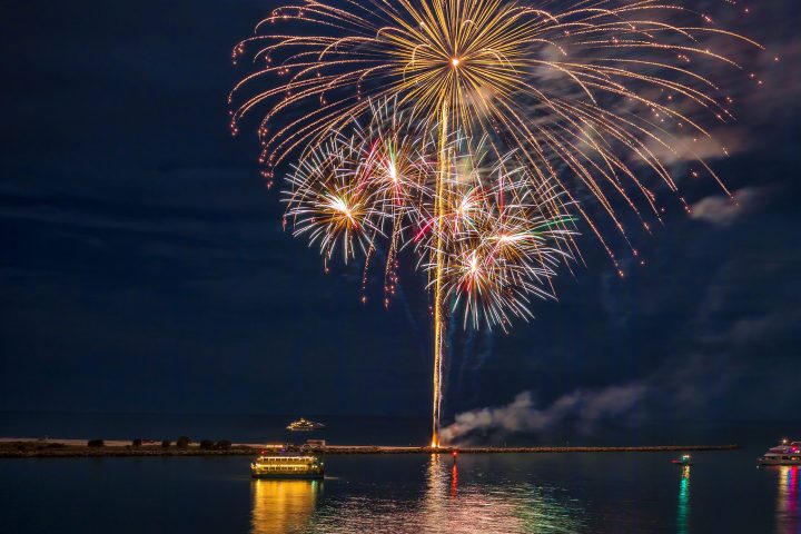 Fireworks over a calm waterfront seen from a sandy beach with lights and people watching. photo by Michael Leonard