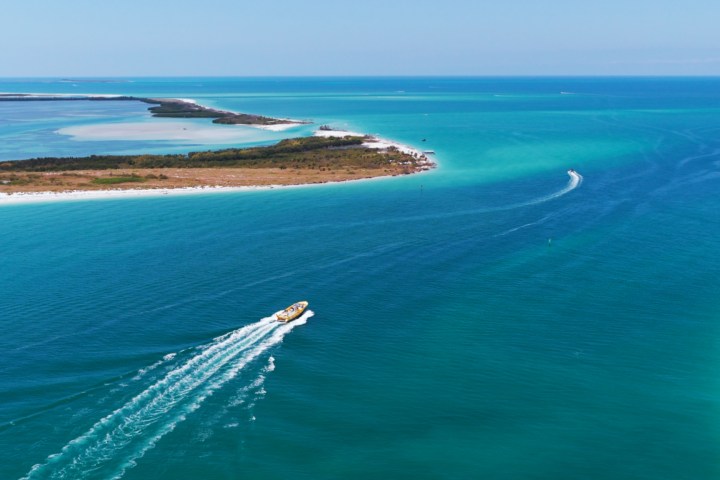 Aerial view of a motorboat leaving a wake in turquoise ocean near a sandy island.