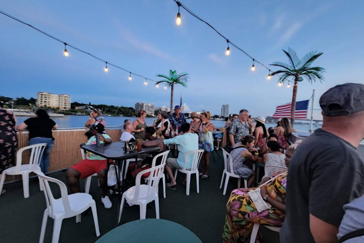 People sitting and standing around tables on a deck with string lights, an artificial palm tree, and a USA flag.