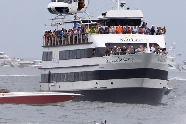 Helicopter over a crowd-filled yacht, with a racing speedboat passing by on the water.