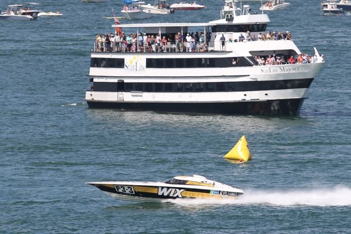 Speedboat racing near a large cruise ship with onlookers, on a sunny day.