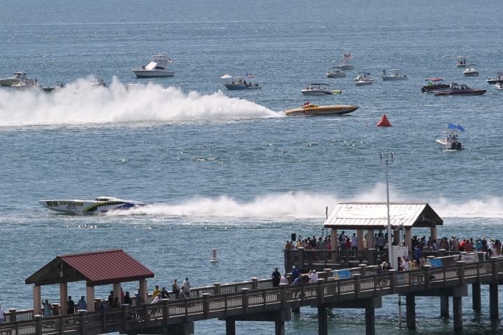 Speedboats racing on water with spectators on a pier, smaller boats in background.