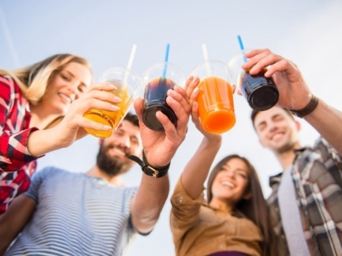 Four smiling people raising plastic cups with colorful drinks outside against a blue sky.