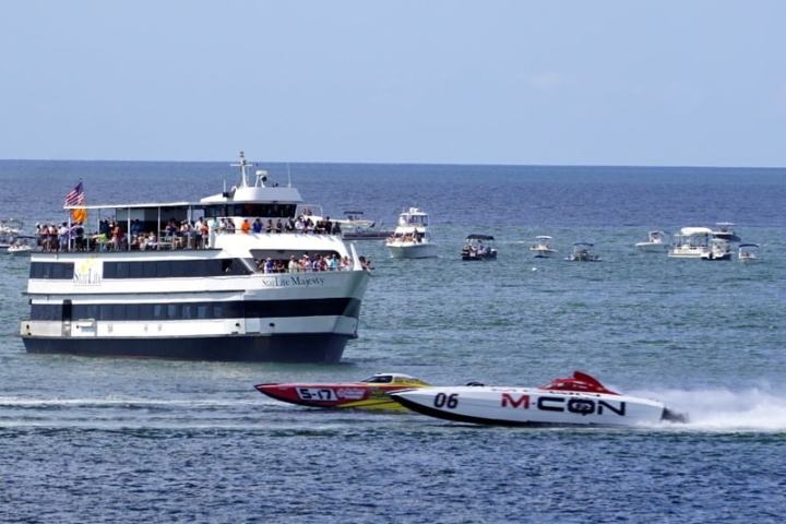 Speedboat races past a large passenger boat on a sunny sea with distant boats visible.