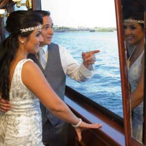 Bride and groom on a boat, groom pointing out the window, bride reflected in glass.