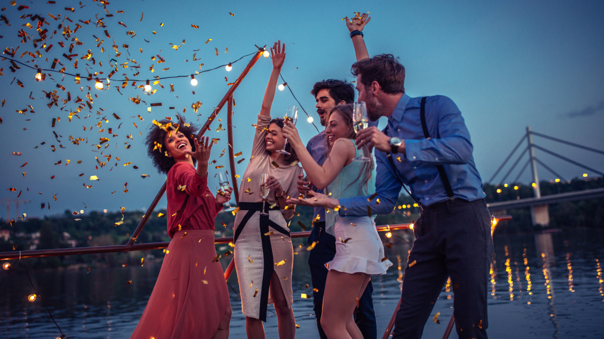Five people celebrating on a boat with lights and confetti in the evening.