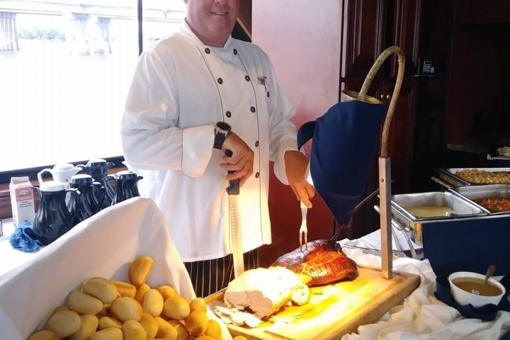Chef in uniform carving meat at a buffet table with bread rolls.