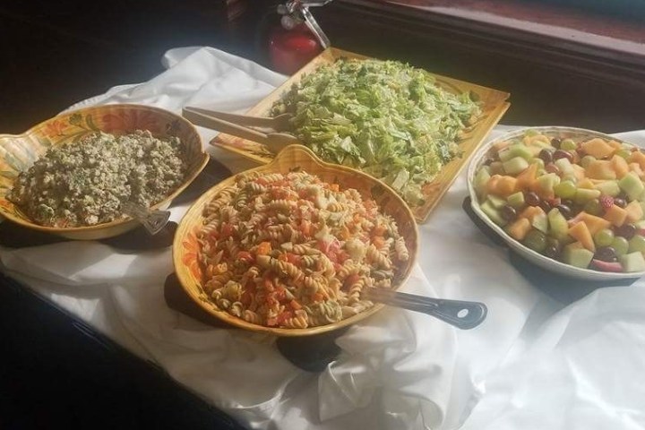 Four bowls of various salads including pasta, greens, fruit, and grain on a table with a white cloth.