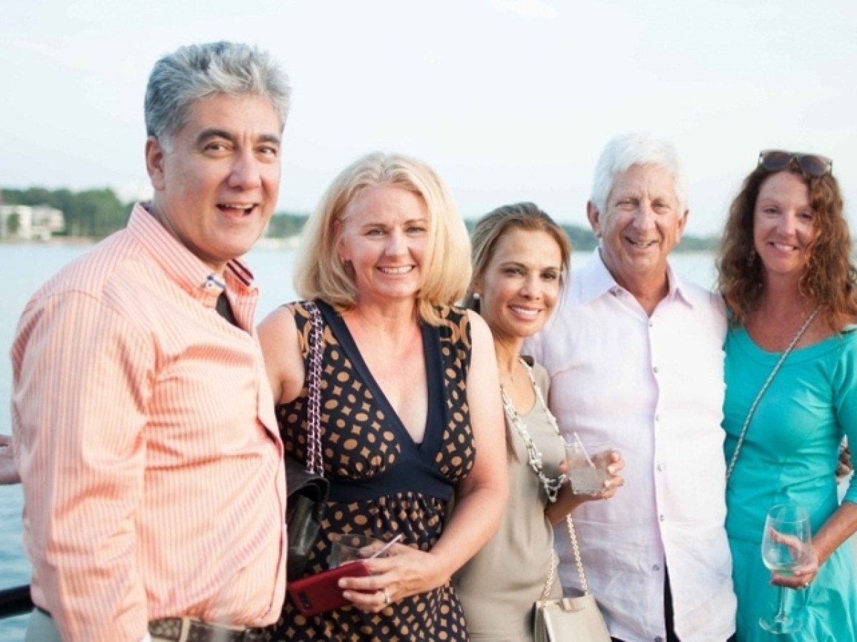 Five people smiling and holding drinks, posing in front of a lake or waterfront.