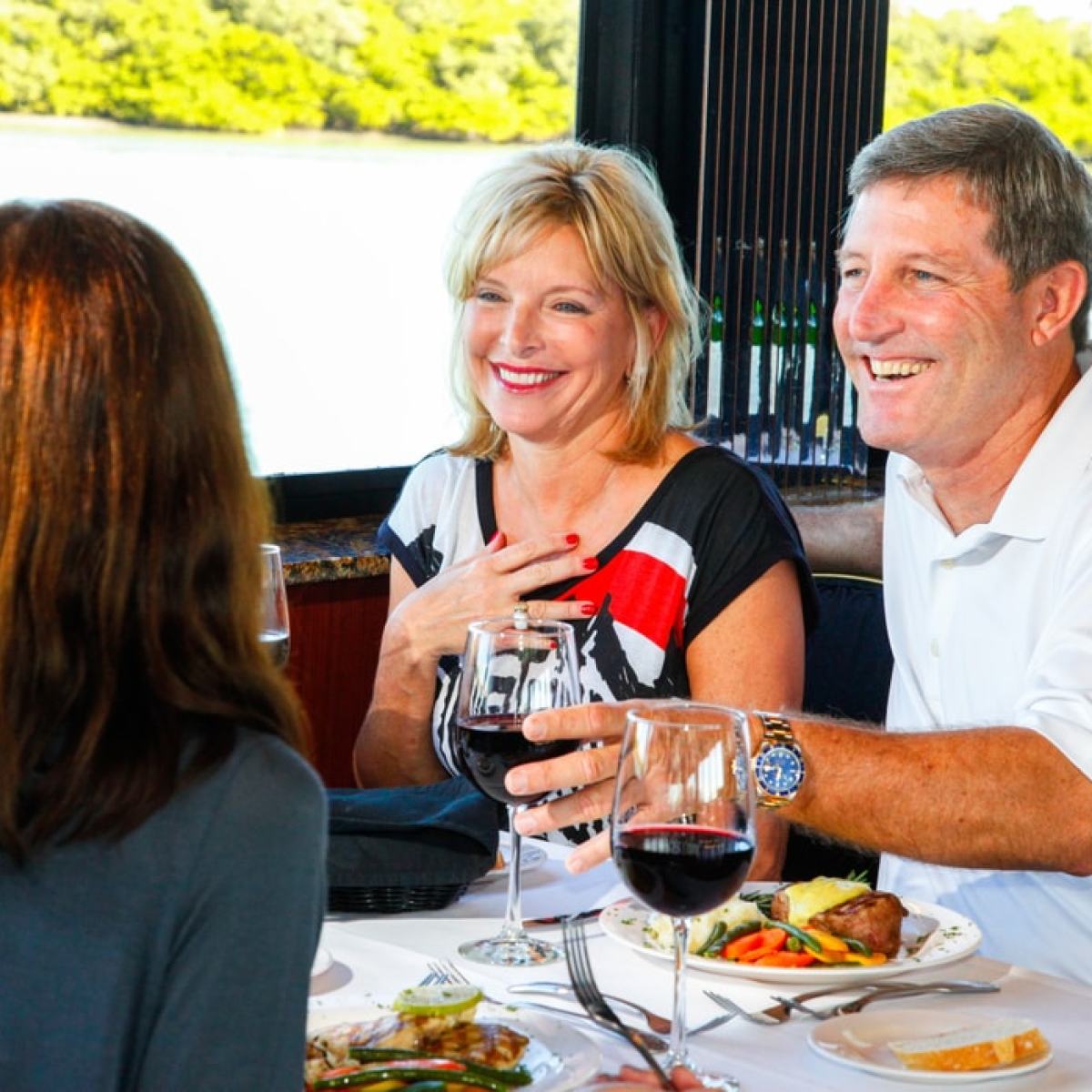 Three people enjoying a meal with wine by a window with a view of greenery outside.