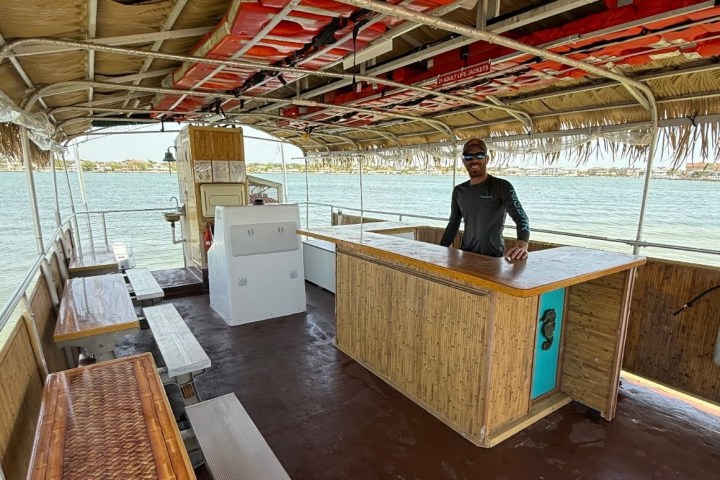 Man on a boat with bamboo bar, canopy, benches, and life jackets overhead, docked by water.