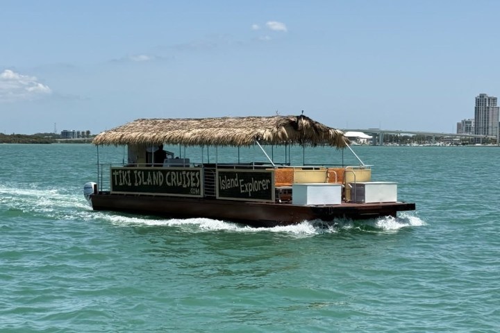 Tiki hut boat on water, labeled 'Tiki Island Cruises', with cityscape in background.