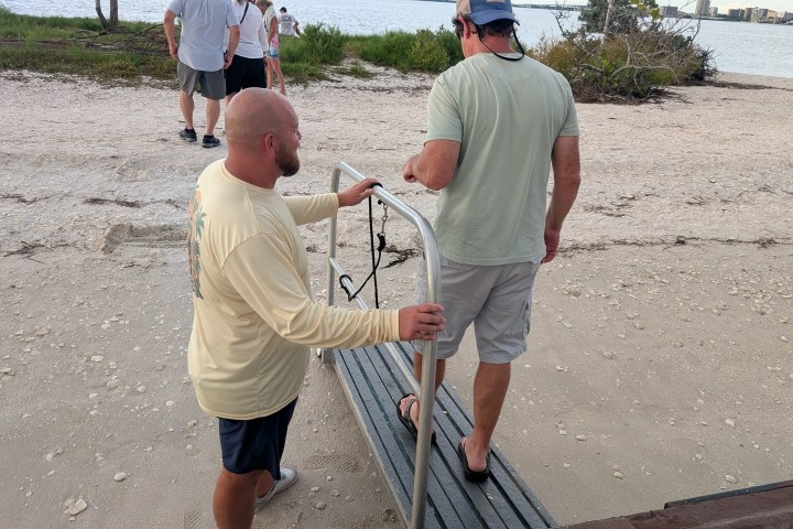 Two men holding a metal railing on a sandy beach pathway, with the sea and shrubs in the background.