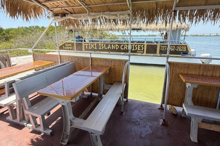 Open-air boat with benches and tables, thatched roof, near water with Tiki Island Cruises sign.