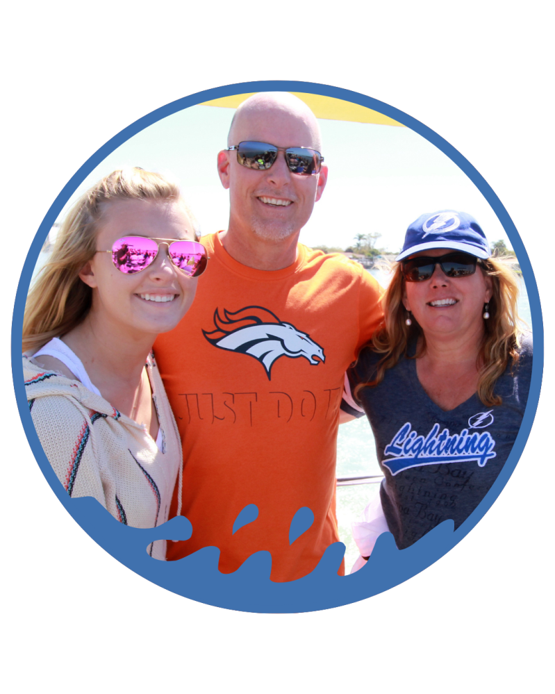 Three people in sunglasses, one wearing an orange shirt, posing outdoors with smiles.