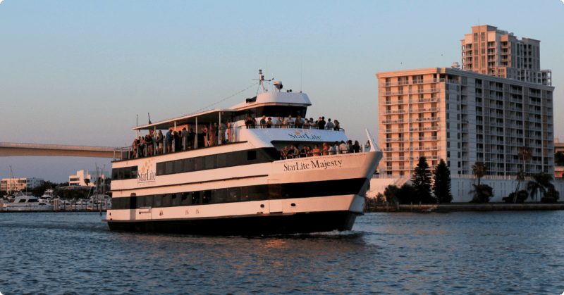 Tour boat with people on the deck cruising near waterfront buildings at sunset.