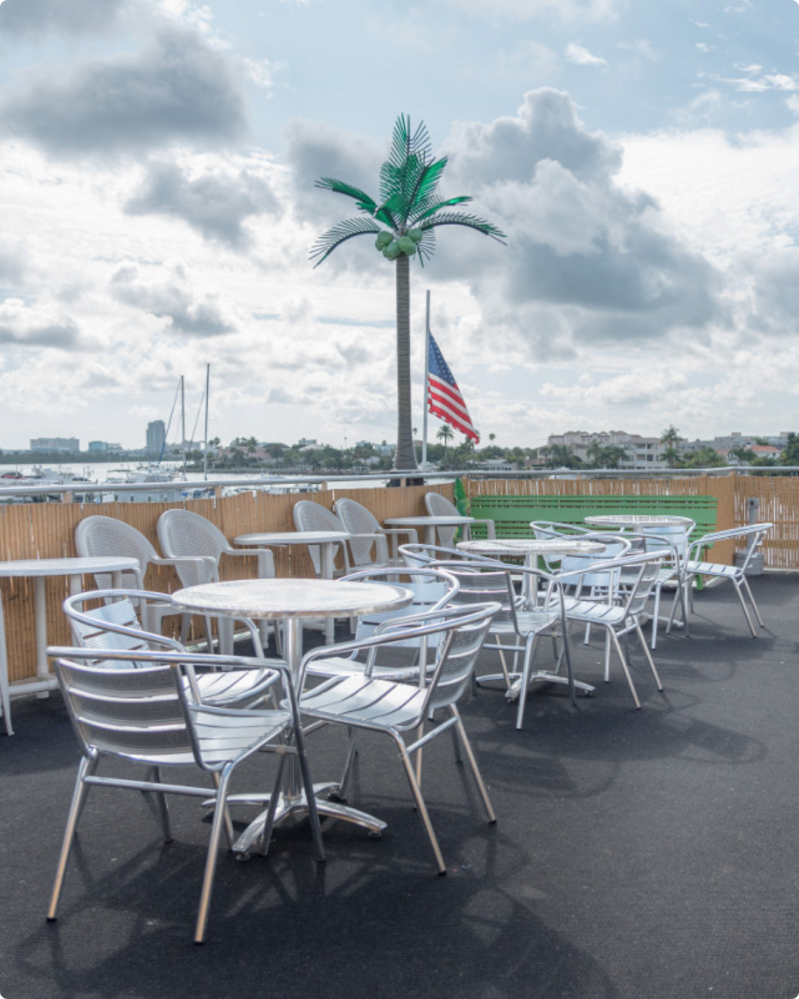 Outdoor patio with metal tables and chairs, artificial palm tree, and a U.S. flag with cloudy sky.