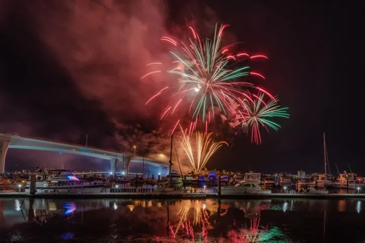 Fireworks over a marina at night, with boats and a bridge in the background.