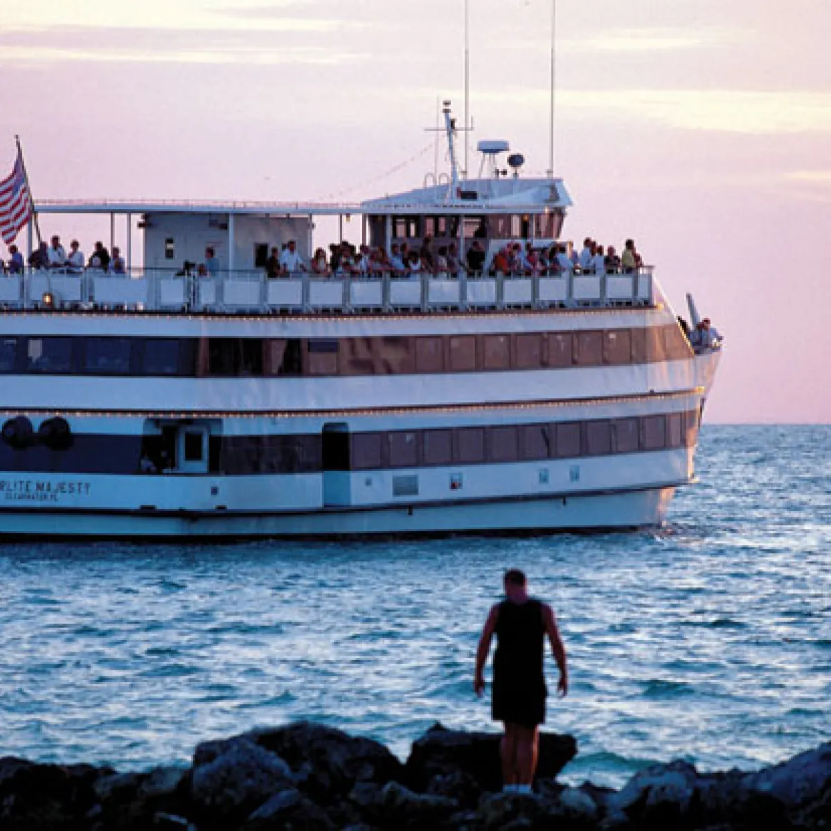 Large vessel with people on deck sails near rocky shore at sunset.