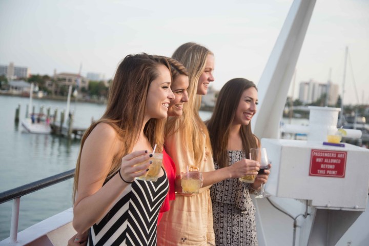 Four women smiling with drinks on a boat, overlooking a marina.
