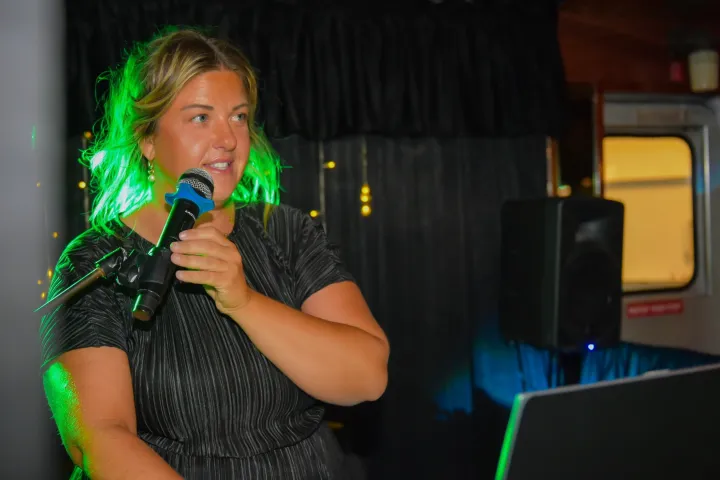 Person speaking into a microphone, standing near a laptop in a dimly lit room.