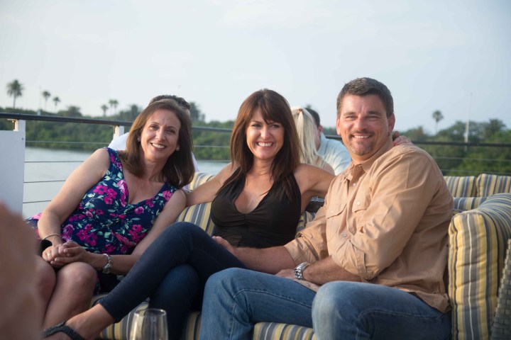 Three people sitting on a couch outside, smiling, with water in the background.