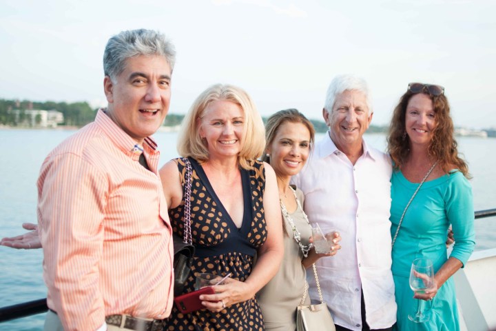 Group of five adults smiling on a boat deck by the water.