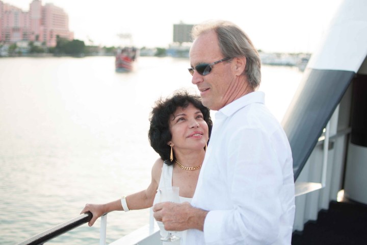 Couple on boat, woman looking up, man wearing sunglasses, holding glass, water and buildings in background.