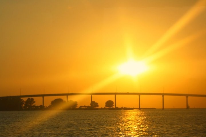 Sunset over bridge with bright sun and water in foreground.