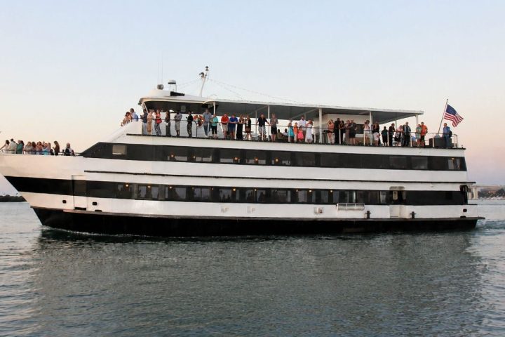Large boat with people on deck cruising on water, flag at stern.
