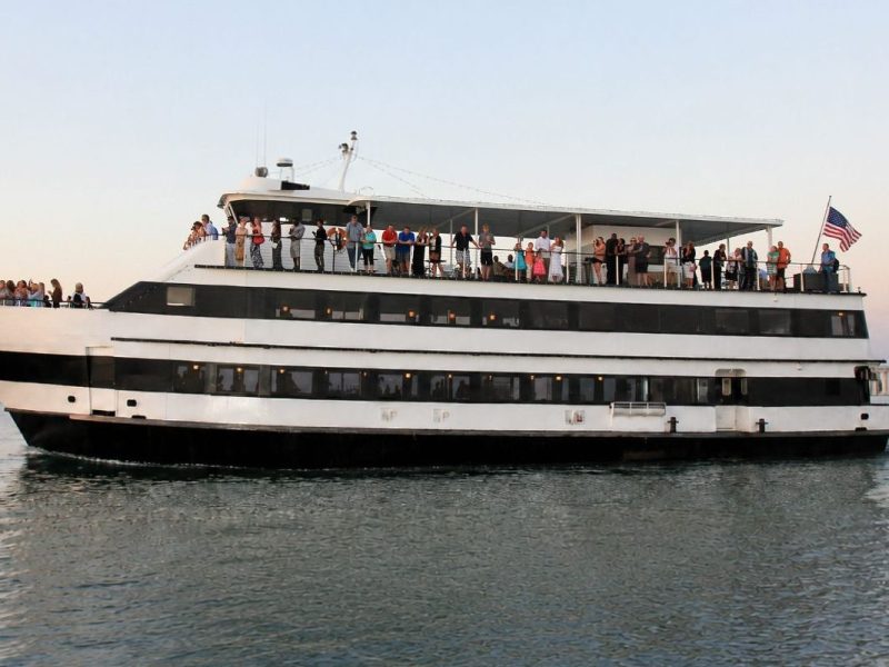 Large boat with people on deck cruising on water, flag at stern.