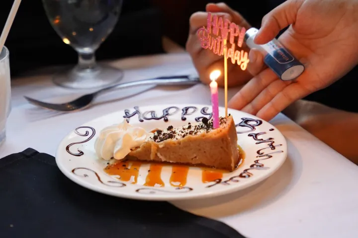 Cake slice with lit candle and 'Happy Birthday' decoration on plate.