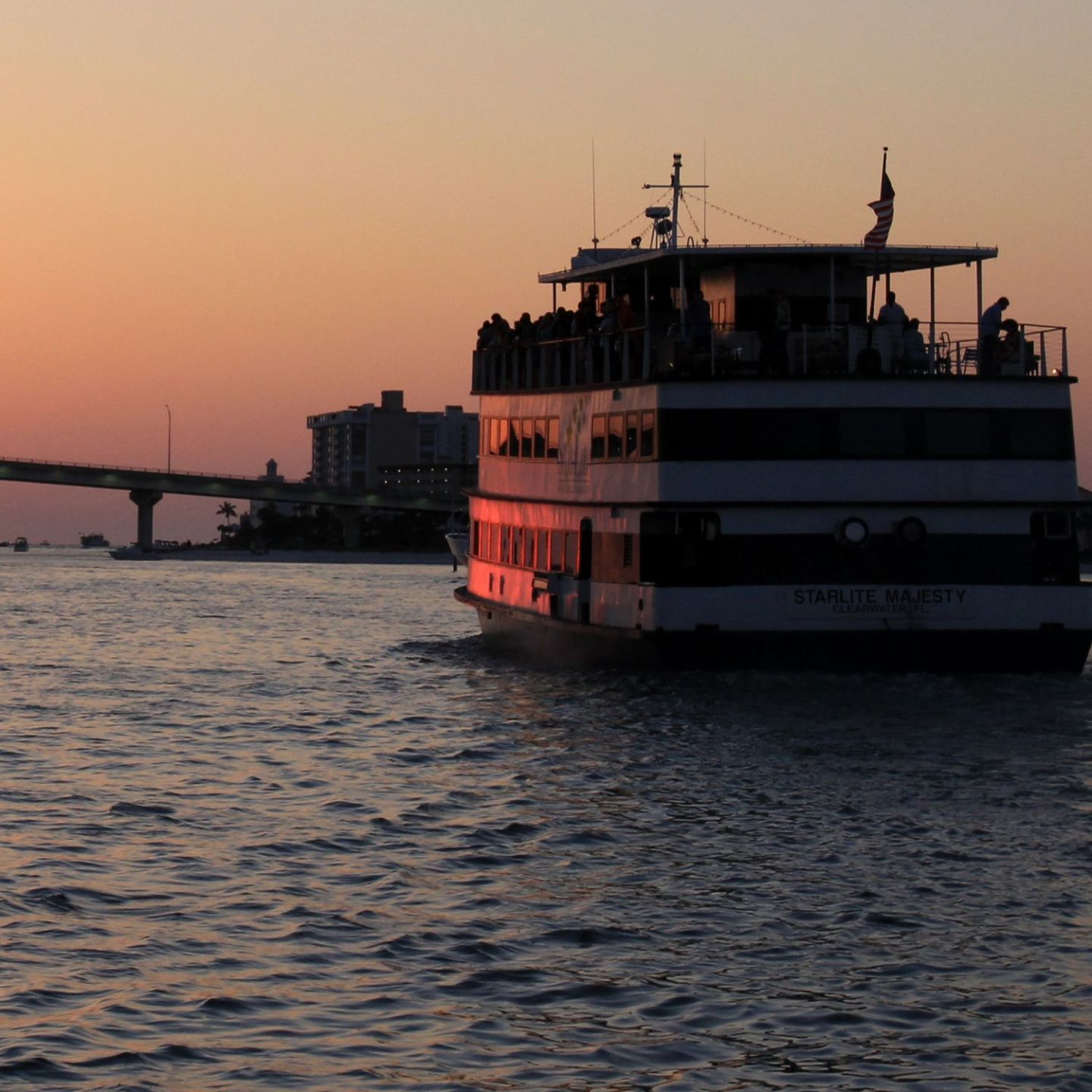 A ferry boat sails at sunset near a bridge, silhouette against the orange sky.