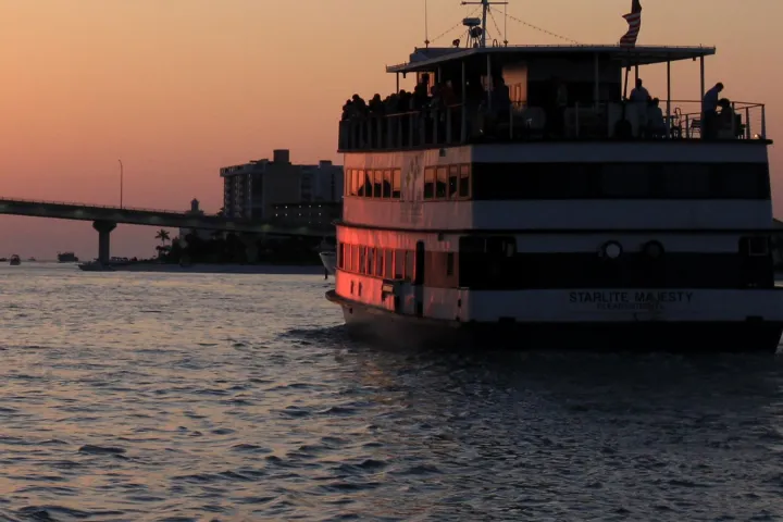 A ferry boat sails at sunset near a bridge, silhouette against the orange sky.