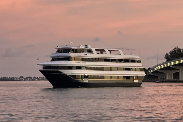 A large boat sails under a bridge at sunset on a calm body of water.