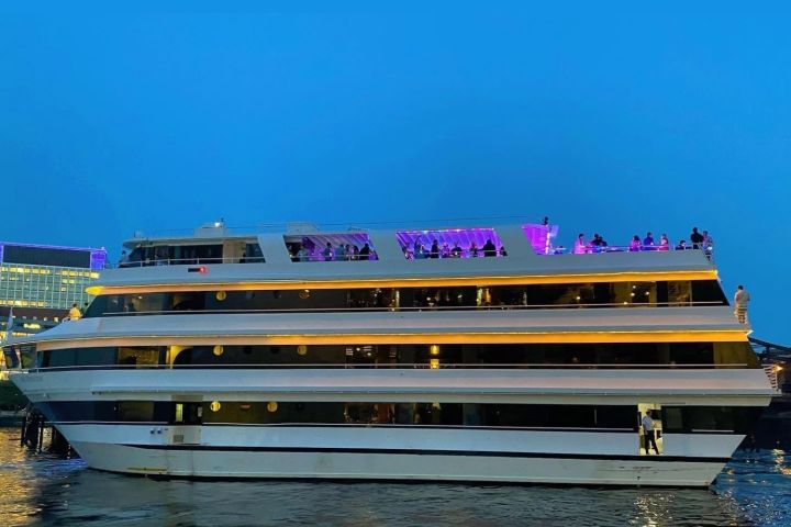 Large white yacht with people on deck, illuminated by purple lights against a twilight sky.