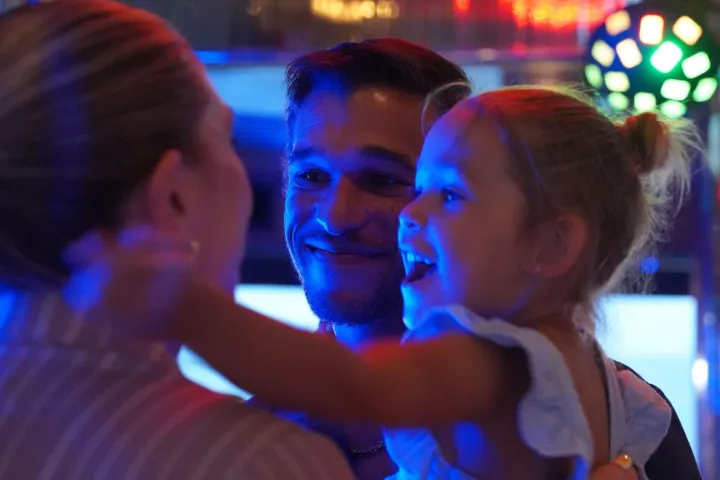Smiling man and woman hold joyful child in a dimly lit room.