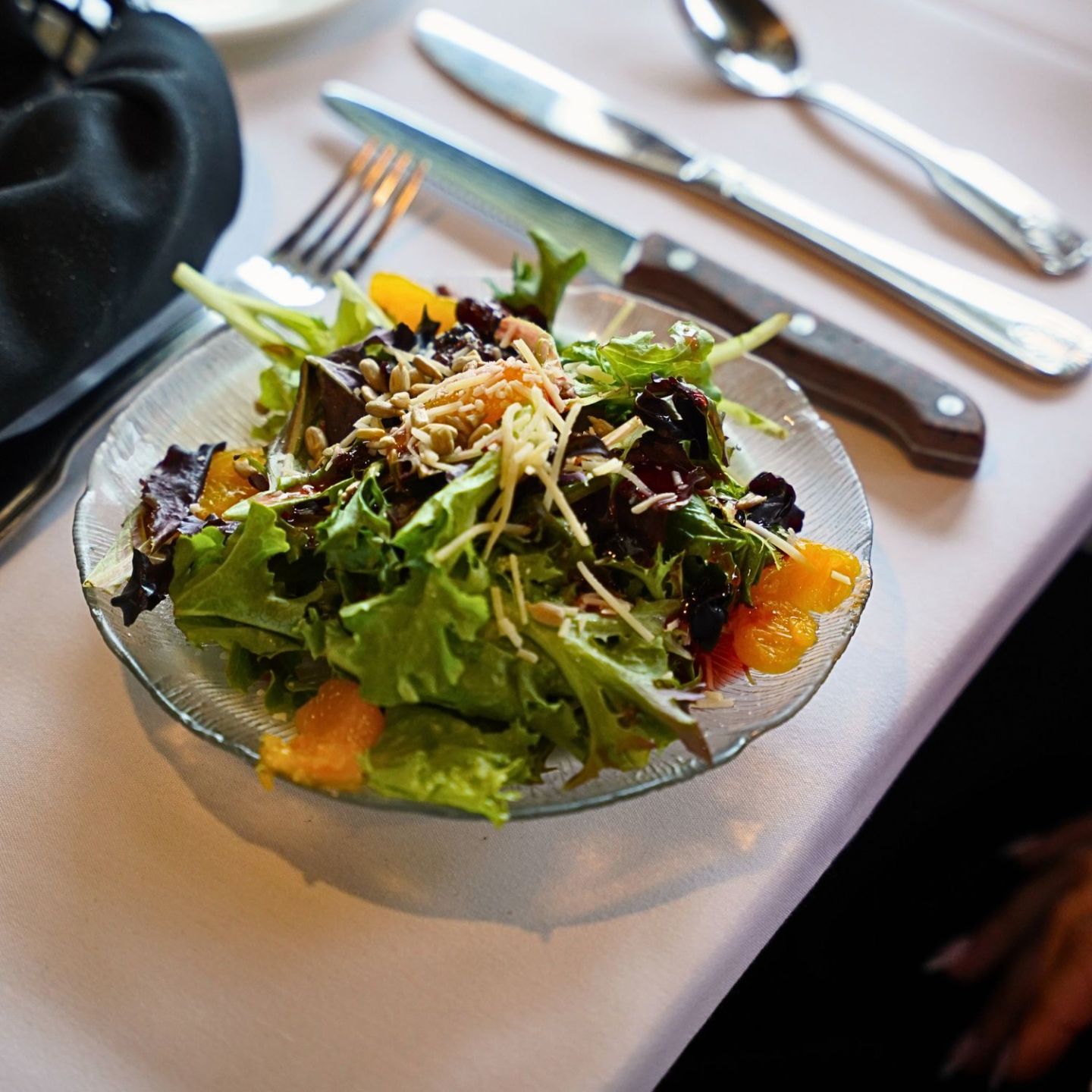 Fresh salad with mixed greens, orange slices, and seeds on a plate, beside cutlery on a table.