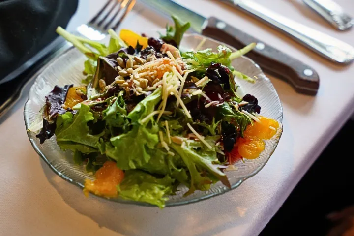 Fresh salad with mixed greens, orange slices, and seeds on a plate, beside cutlery on a table.