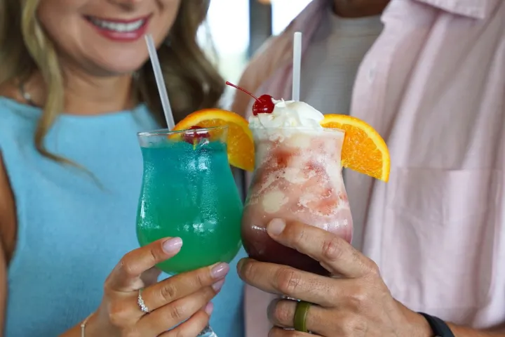 Two people clinking colorful cocktails with fruit garnishes, smiling.