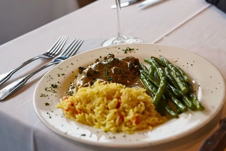 Plate with rice, green beans, meat, beside a glass of red wine on a table.