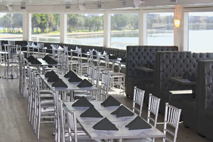 Dining area with silver chairs, black booths, and tables set with black napkins, overlooking a waterfront view.