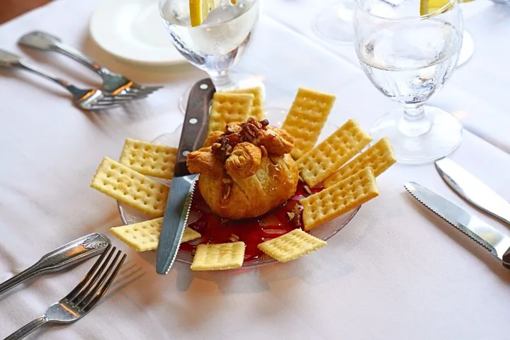 Cheese ball with crackers and knife on a plate, surrounded by water glasses with lemon.
