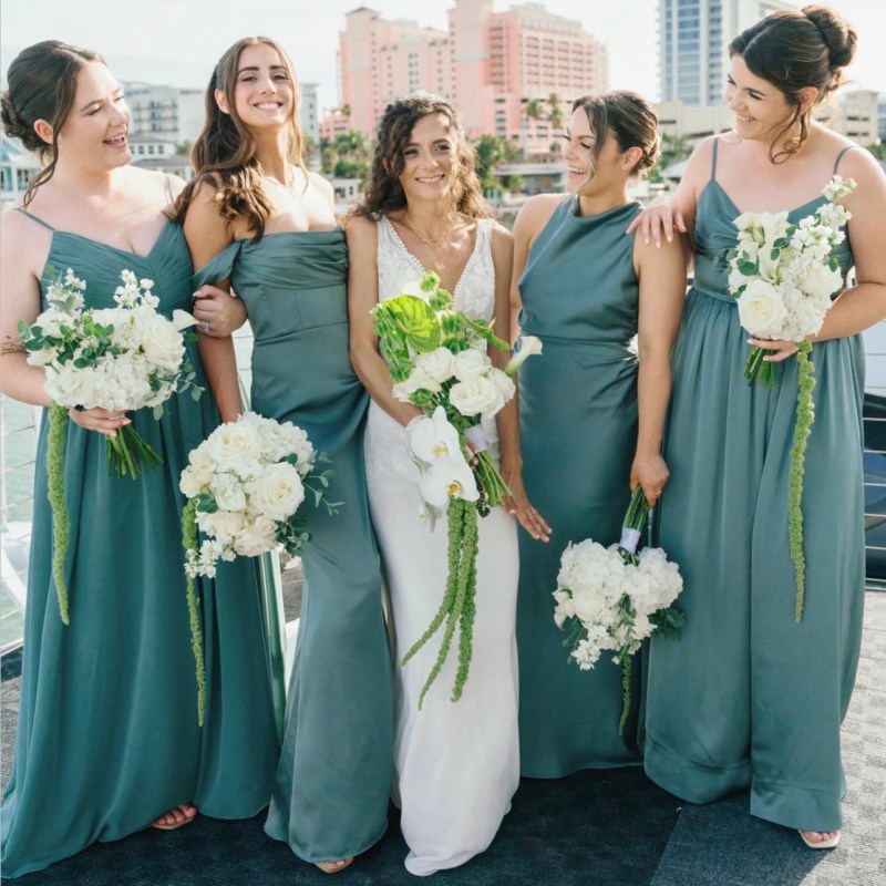 Bride with four bridesmaids in teal dresses holding white bouquets, smiling outdoors.