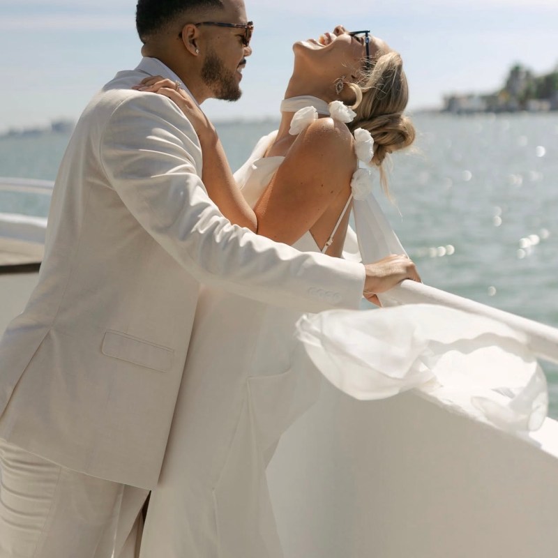 Couple in white attire laughing on a boat, with water and skyline in background.