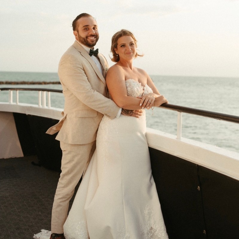 Couple in wedding attire on a boat deck, smiling at sunset.