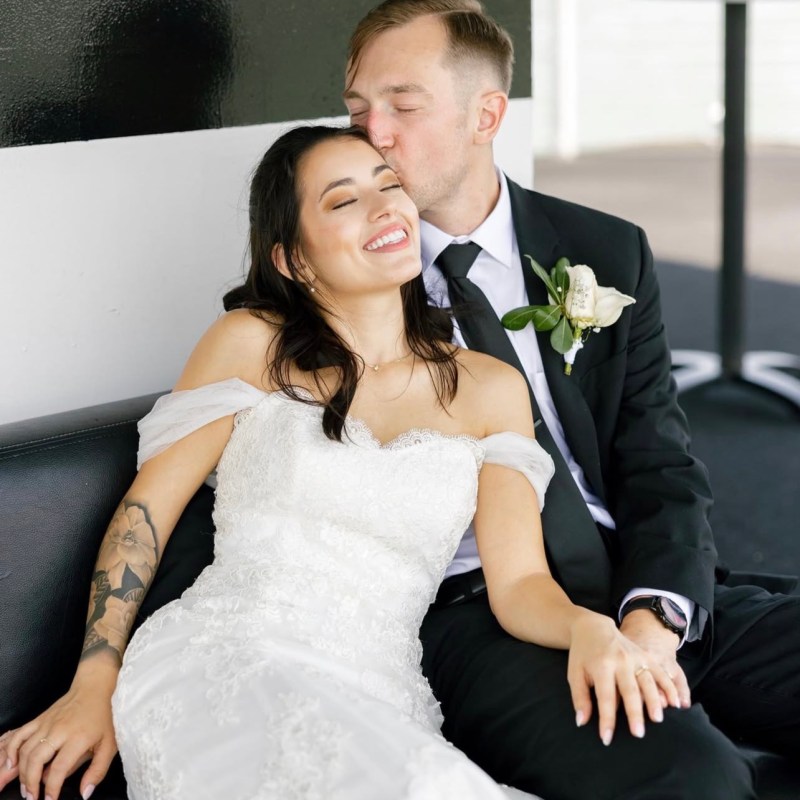Smiling bride and groom sitting, groom kissing bride's forehead.