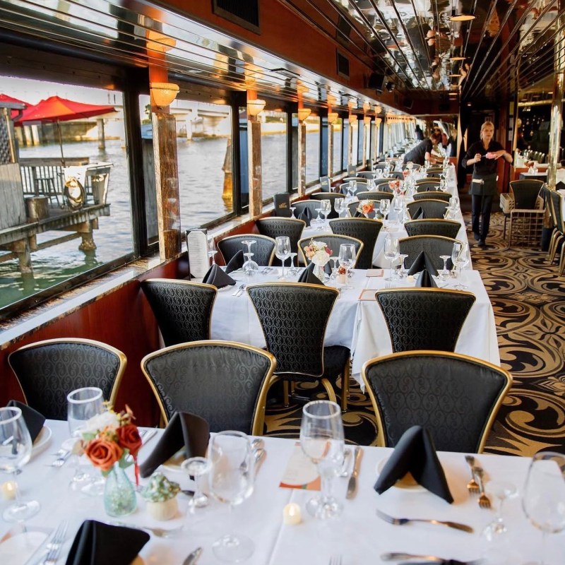 Elegant dining setup on a boat with white tablecloths, black napkins, and water view.