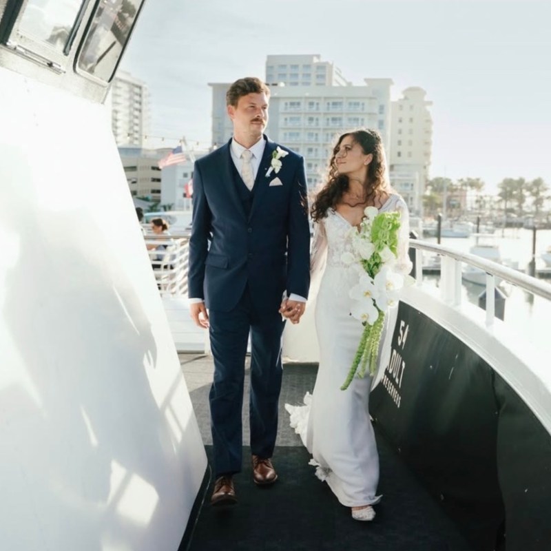 Bride and groom holding hands on a boat, city skyline in background.
