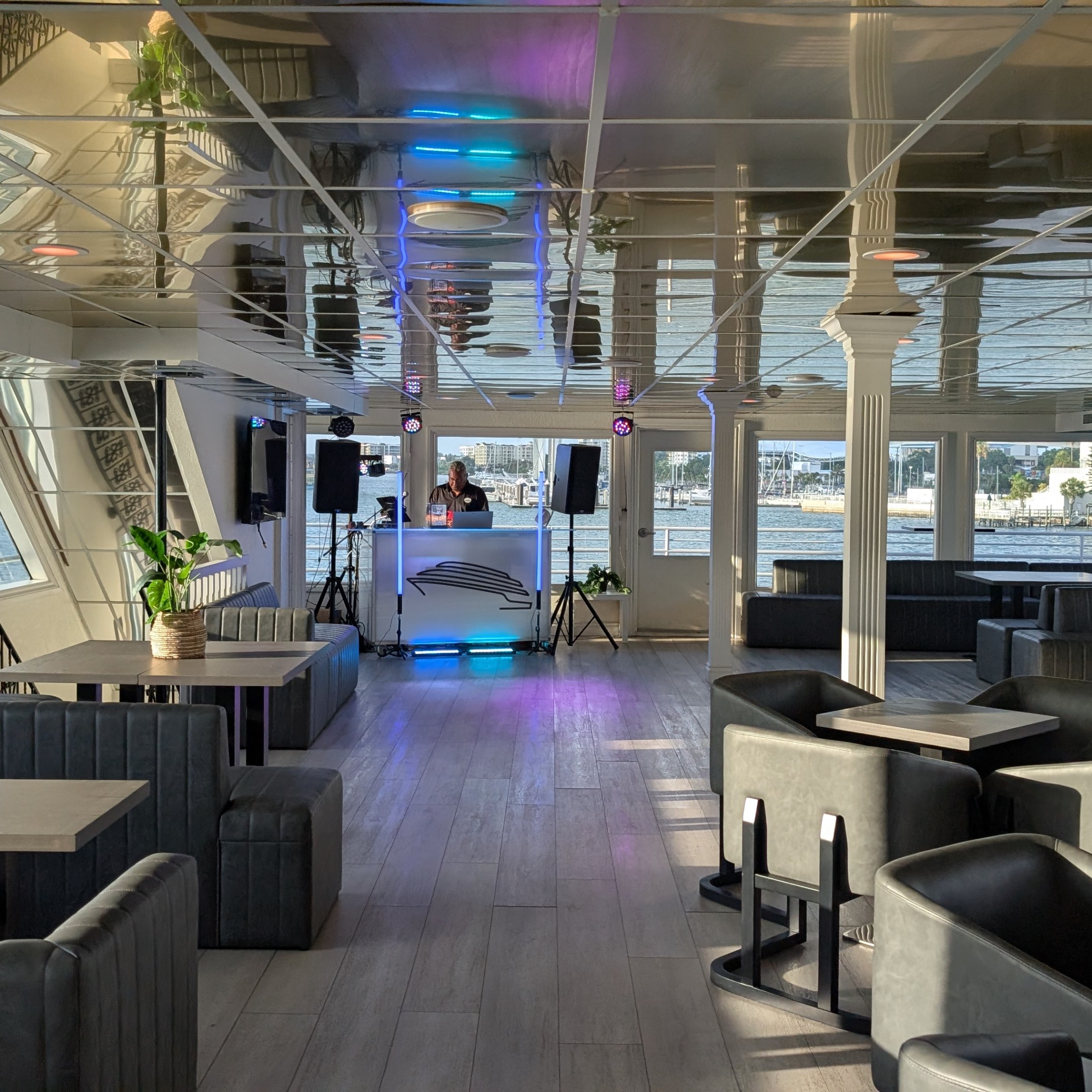 Spacious, empty modern boat lounge with tables, black seating, and a DJ setup near large windows.